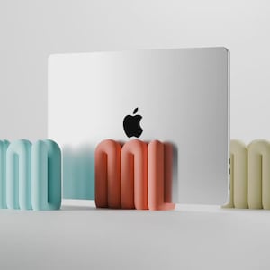 Op de afbeelding: Een zilveren laptop met een zwart Apple-logo wordt rechtop gehouden door drie afzonderlijke, U-vormige, afgeronde standaards in pastelkleuren. De linker standaard is lichtblauw, de middelste koraalkleurig en de rechter is lichtgeel, alle glad.
