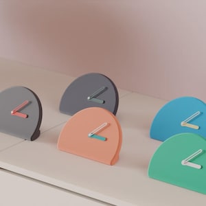 Peut inclure: Cinq horloges de bureau semi-circulaires de différentes couleurs, notamment gris foncé, gris clair, pêche, bleu et vert. Chaque horloge présente une base plate, un sommet incurvé et deux aiguilles contrastées fixées au centre du cadran.