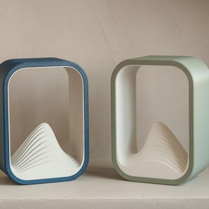 Puede incluir: Dos l&aacute;mparas decorativas modernas. Una es azul, la otra verde. Cada l&aacute;mpara tiene un marco cuadrado con esquinas redondeadas y un dise&ntilde;o blanco en capas en forma de monta&ntilde;a en el interior. Las l&aacute;mparas est&aacute;n sobre una superficie blanca.