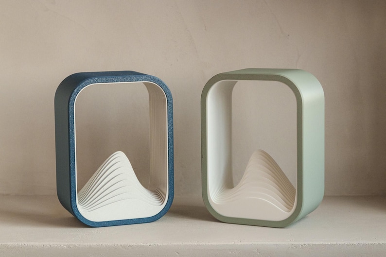 Puede incluir: Dos l&aacute;mparas decorativas modernas. Una es azul, la otra verde. Cada l&aacute;mpara tiene un marco cuadrado con esquinas redondeadas y un dise&ntilde;o blanco en capas en forma de monta&ntilde;a en el interior. Las l&aacute;mparas est&aacute;n sobre una superficie blanca.