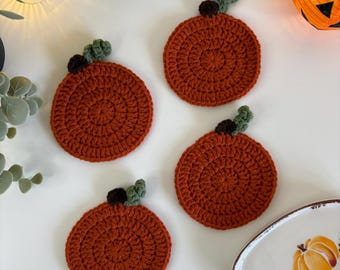 Pode incluir: Quatro bases para copos redondas de crochê laranja em forma de abóbora, com detalhes verdes e castanhos. Estão dispostas sobre uma superfície branca, com um prato decorativo com um desenho de abóbora. Tema outonal.