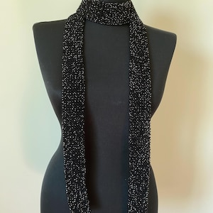 May include: A black scarf with a subtle sparkle effect, displayed on a mannequin. The scarf is long and narrow, with a textured appearance. The fabric appears to be woven or knitted, with small, reflective elements throughout.