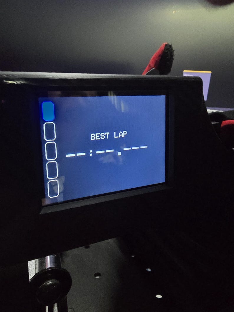 Pu&ograve; includere: Uno schermo digitale nero montato in un alloggiamento scuro. Lo schermo mostra il testo BEST LAP in bianco, seguito da una linea tratteggiata per il timer. A sinistra appare una colonna verticale di cinque icone rettangolari, con l'icona superiore illuminata in blu.