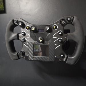 May include: A black carbon fiber racing steering wheel featuring a central rectangular display screen, multiple circular buttons with white icons, and yellow-tipped rotary knobs. The device has ergonomic side grips and a flat-bottom design.
