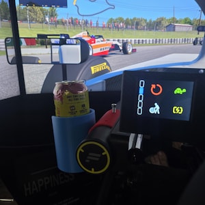 Puede incluir: Un simulador de carreras con un volante que tiene una pantalla digital con iconos de parada en boxes, tortuga y energía. Una lata de bebida energética amarilla está en un portavasos azul claro, frente a una pantalla de juego de carreras.