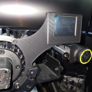 May include: A black racing steering wheel attachment featuring a carbon fiber texture, a rectangular digital display screen, and a power button. The device includes a circular central hub with visible screws and a side-mounted control knob with a yellow ring.