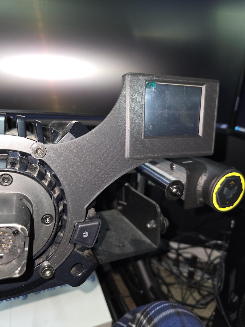 Pu&ograve; includere: Un accessorio per volante da corsa nero con trama in fibra di carbonio, uno schermo digitale rettangolare e un pulsante di accensione. Il dispositivo include un mozzo centrale circolare con viti a vista e una manopola di controllo laterale con un anello giallo.