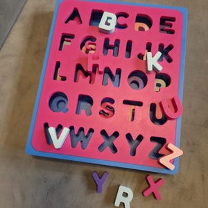 May include: A wooden alphabet puzzle with a pink central section, a blue border, and cut-out letter shapes. Several individual letters in white, purple, and pink are scattered around the board. The letters are designed to fit into the corresponding cut-out spaces.
