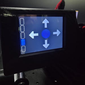 Pu&ograve; includere: Un pannello di controllo elettronico nero con uno schermo digitale rettangolare. Il display mostra un'icona circolare blu al centro circondata da quattro frecce direzionali bianche rivolte verso l'alto, il basso, sinistra e destra. A sinistra si trova una colonna verticale di cinque pulsanti rettangolari, con il quarto pulsante evidenziato in blu.