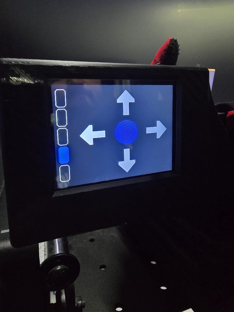 Pu&ograve; includere: Un pannello di controllo elettronico nero con uno schermo digitale rettangolare. Il display mostra un'icona circolare blu al centro circondata da quattro frecce direzionali bianche rivolte verso l'alto, il basso, sinistra e destra. A sinistra si trova una colonna verticale di cinque pulsanti rettangolari, con il quarto pulsante evidenziato in blu.