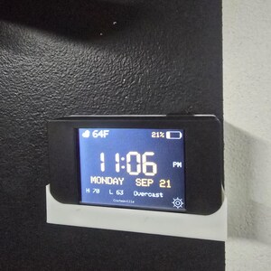 Puede incluir: Un dispositivo de pantalla digital negro montado en un soporte blanco, que muestra la hora 11:06 PM, la fecha lunes 21 de septiembre, una temperatura de 64F, 21% de batería y condiciones climáticas nubladas en Coatesville con un icono de sol.