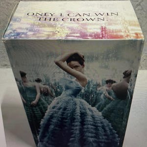 May include: A box with the text "ONLY I CAN WIN THE CROWN." The box features a woman in a blue ruffled gown, with other figures in similar gowns in the background. The overall aesthetic is artistic and elegant.