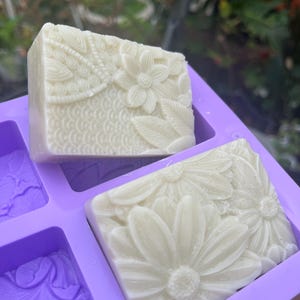 Puede incluir: Dos barras rectangulares de jabón blanco con diseños florales se muestran en un molde morado. Una barra presenta un patrón de flores y hojas, mientras que la otra tiene un diseño de margaritas. El jabón es sin perfume y hecho a mano.