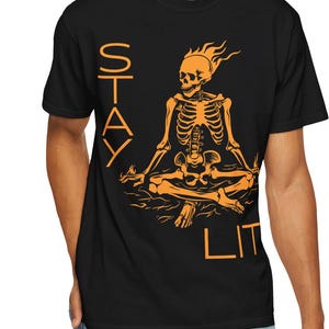 May include: Black t-shirt with an orange graphic of a skeleton in a meditative pose, with flames above its head. The words "STAY LIT" are printed vertically on the left and bottom right of the design.