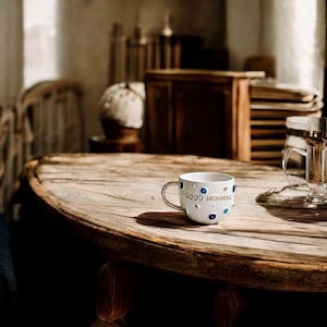 May include: A white ceramic mug with a metallic handle featuring the text GOOD MORNING in gold lettering. The exterior is decorated with small, raised blue and white circular eye-shaped accents and silver dots scattered across the surface.