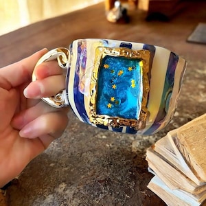 Puede incluir: Una taza de cerámica con rayas verticales azules y blancas y acabado iridiscente. La taza tiene un asa dorada y un centro rectangular con marco dorado que contiene una incrustación de resina azul decorada con pequeñas estrellas amarillas.