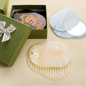 May include: An open green gift box with a white ribbon bow next to a tortoiseshell comb with the name "KATIE" in gold lettering. A light blue comb and mirror set is also visible, along with a white comb with the name "OLIVIA" in gold lettering.