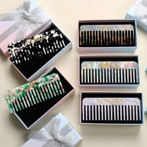 May include: Several personalized combs in gift boxes. One comb is black and white, another is green and white, and others are pearlescent. Each comb has a name in gold lettering. The combs are in white boxes with black interiors, and some are decorated with silver ribbon.
