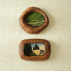 May include: Two small, rustic wooden frames with landscape paintings. The top frame is oval, the bottom is rectangular. The paintings depict a green field and a building with trees. The frames are a warm brown colour.