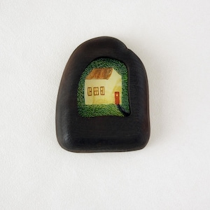 May include: A dark brown, pebble-shaped wooden brooch. The brooch features a small, detailed painting of a house with a brown roof and red door, set within a green, grassy area. The house has three windows.