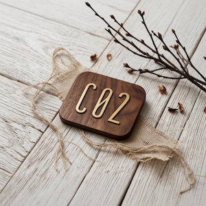 May include: A square wooden plaque with the letters "CO2" in gold-colored metal. The plaque rests on a piece of burlap fabric, with a twig and small buds in the background. The wooden surface is light-colored.