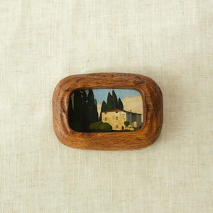 May include: A small, rectangular wooden frame with rounded edges, containing a painting of a house and trees. The painting features a light-colored building with a blue sky and tall, dark green trees. The frame is a warm brown color.