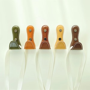 May include: Five leather towel hooks in green, orange, brown, yellow, and dark brown. Each hook has a loop and is attached with two silver rivets. White towels hang from the hooks.