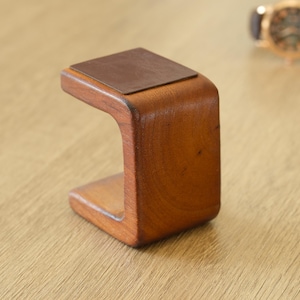 May include: A wooden watch stand with a curved design. The stand is made of warm-toned wood and has a rectangular, leather-like pad on top. A gold-toned watch with a brown leather strap is slightly out of focus in the background.