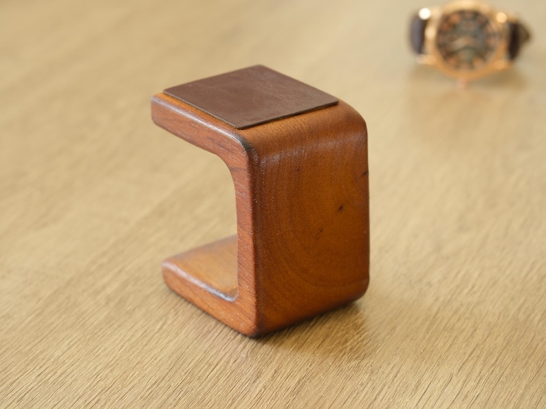 May include: A wooden watch stand with a curved design. The stand is made of warm-toned wood and has a rectangular, leather-like pad on top. A gold-toned watch with a brown leather strap is slightly out of focus in the background.