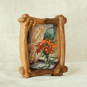 Peut inclure: Tableau encadré représentant des lys orange dans un vase en verre près d'une fenêtre avec un rideau à motifs floraux. Le cadre en bois brun clair présente un design rustique. La peinture est de style aquarelle.
