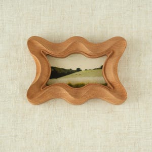 May include: A small, wooden picture frame with a wavy, ornate design. The frame is made of light brown wood and holds a landscape image of a green field and hills. The frame is approximately 6 inches wide and 4 inches tall.