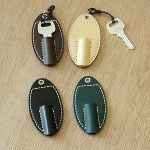 May include: Four leather key holders in brown, beige, black, and green. Each oval-shaped holder has a stitched white border and a small pocket for a key. One holder has a key inserted.