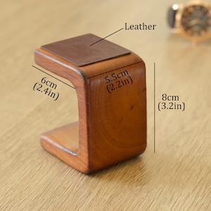 May include: A wooden watch stand with a leather top. The stand is 6cm wide, 5.5cm deep, and 8cm tall. The stand is a warm brown colour and has a curved design. A watch is visible in the background.