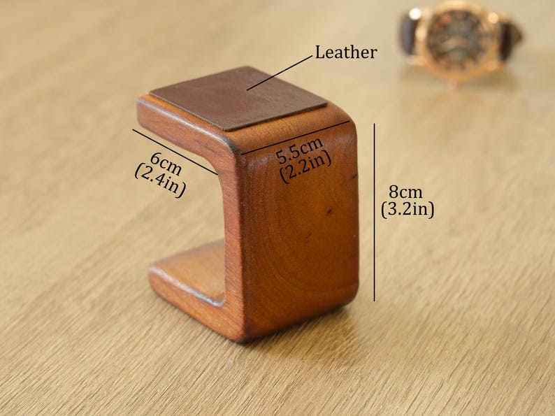 May include: A wooden watch stand with a leather top. The stand is 6cm wide, 5.5cm deep, and 8cm tall. The stand is a warm brown colour and has a curved design. A watch is visible in the background.