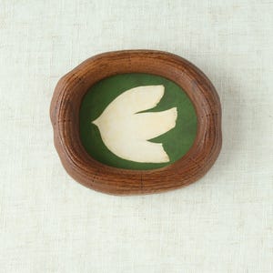 May include: A wooden framed artwork featuring a white dove against a green background. The frame is oval-shaped with a textured, brown finish. The dove is stylized with three distinct wing shapes.
