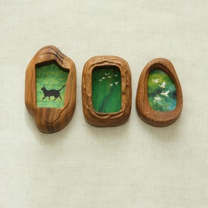 May include: Three wooden wall art pieces with irregular shapes. Each piece contains a miniature painting. One shows a black cat, another has white birds, and the third depicts birds in flight. The frames are a warm brown colour.