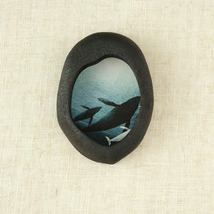 May include: A black, oval wooden frame featuring a whale illustration. The artwork shows whales swimming in a blue ocean. The frame has a natural, organic shape, and the background is a light beige.