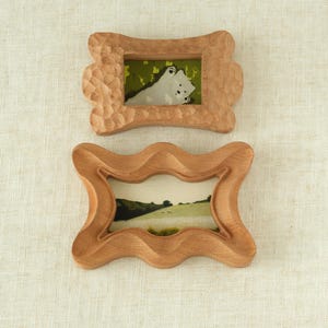 May include: Two small, wooden picture frames with a natural finish. The top frame has a carved texture and a picture of a white animal on a green background. The bottom frame has a smooth, wavy design and a landscape photo.