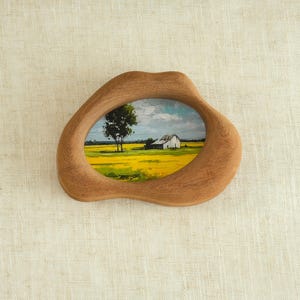 May include: A wooden frame with an organic shape surrounds a small painting of a rural landscape. The painting depicts a yellow field, a white building, a tree, and a blue sky with clouds. The frame is made of light brown wood.