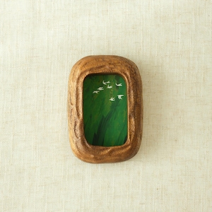 May include: A small, rectangular wooden frame with a natural wood finish. The frame contains a green painted background with white bird silhouettes. The frame is set against a neutral, textured background.