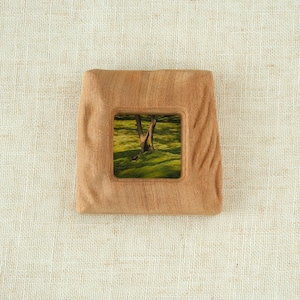 May include: Square wooden frame with a recessed center displaying a landscape scene of a tree in a green field. The frame has a natural wood grain and a slightly textured surface.