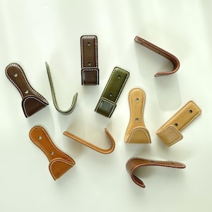 May include: A collection of leather hooks in shades of brown, green, and tan. The hooks are curved and stitched with white thread. Some have a rectangular base, while others have a paddle shape. Measurements are in centimetres.