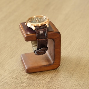 May include: A gold-toned wristwatch with a brown leather band rests on a wooden watch stand. The stand is a dark brown, C-shaped design, showcasing the watch's face and band. The watch has a green face.