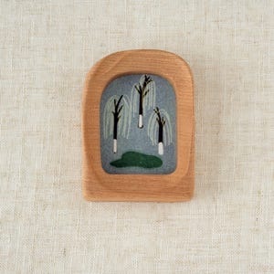 May include: A wooden brooch with a rectangular shape and rounded top. The brooch features a felt insert with a painting of three weeping willow trees with white and black trunks and green foliage. The brooch is set against a neutral background.