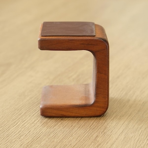 May include: A wooden watch stand with a dark brown leather top. The stand is made of light brown wood and has a curved design. The watch stand is designed to hold a wristwatch.