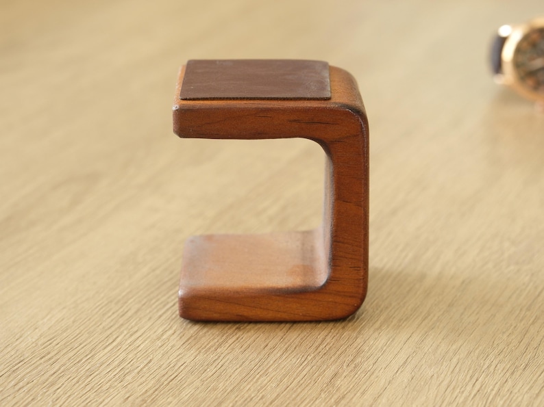 May include: A wooden watch stand with a dark brown leather top. The stand is made of light brown wood and has a curved design. The watch stand is designed to hold a wristwatch.