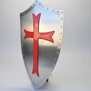 May include: A heater-style shield made of polished silver-toned metal featuring a prominent red cross emblem centered on the front. The shield is bordered by a reinforced metal rim secured with evenly spaced rivets along the entire outer edge.