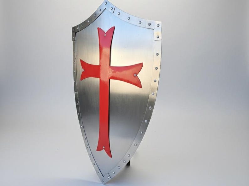 May include: A heater-style shield made of polished silver-toned metal featuring a prominent red cross emblem centered on the front. The shield is bordered by a reinforced metal rim secured with evenly spaced rivets along the entire outer edge.