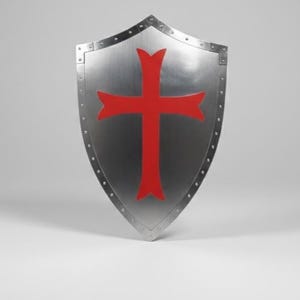 May include: A heater-style shield made of polished silver-toned metal featuring a prominent red cross emblem centered on the front surface. The shield is constructed with a reinforced border secured by a series of visible metal rivets along the edges.