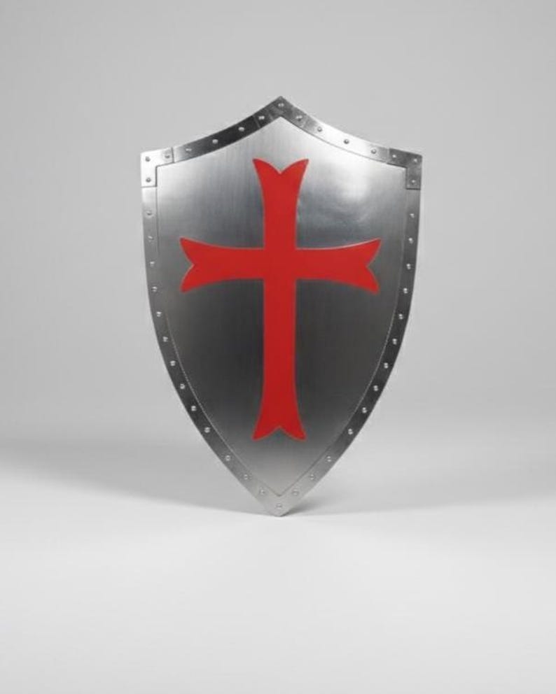 May include: A heater-style shield made of polished silver-toned metal featuring a prominent red cross emblem centered on the front surface. The shield is constructed with a reinforced border secured by a series of visible metal rivets along the edges.